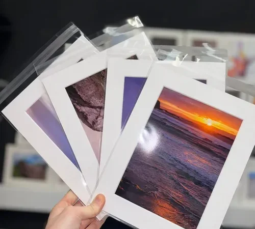 Hand holding mounted photo prints including sunset and landscape photographs