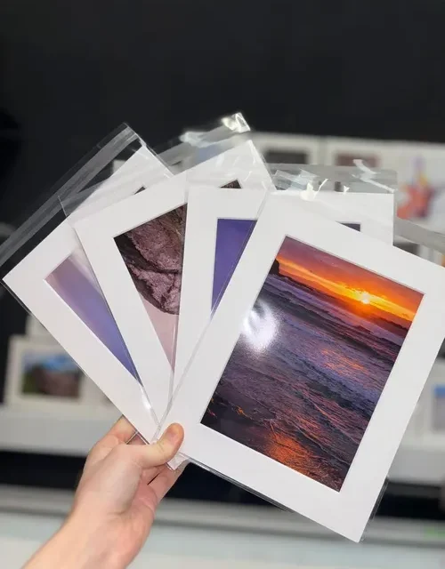 Hand holding mounted photo prints including sunset and landscape photographs
